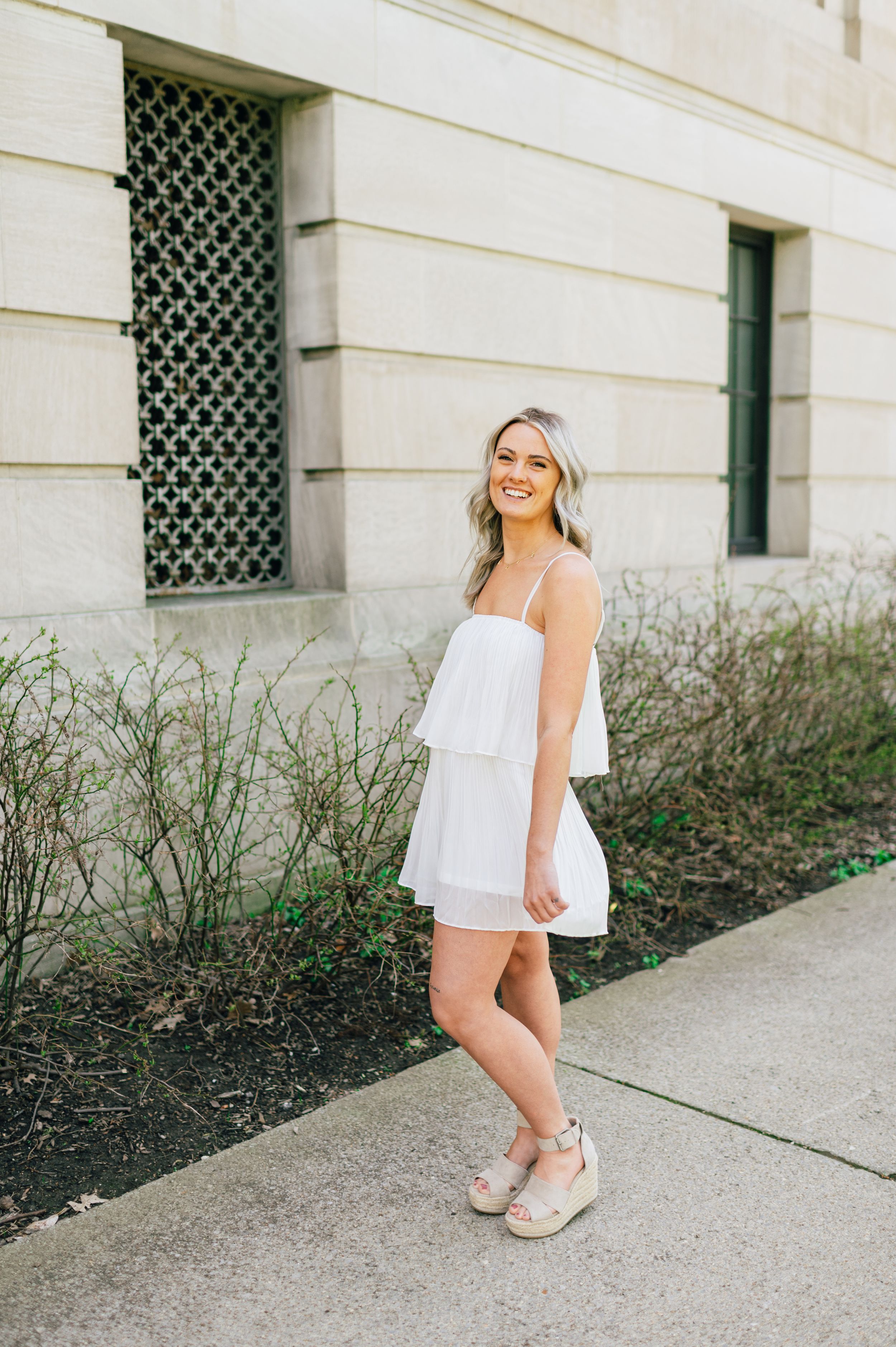 A person in a flowy white summer dress and platform sandals poses on a sidewalk near an ornate building exterior.