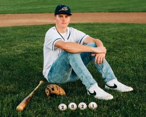 Baseball player sits in grass with baseballs, glove and bat on field while wearing uniform and baseball cap.