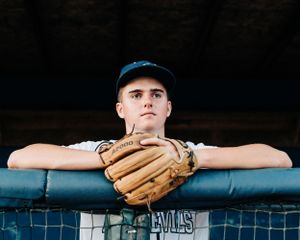 Baseball player wearing blue cap leans on dugout railing with leather glove in focus against dark background.