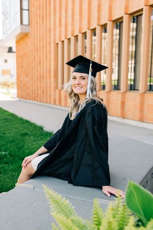 A graduate in traditional black cap and gown sits on a concrete ledge outside a brick building with green grass and plants.