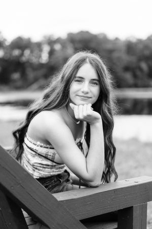 A black and white portrait of a young person with long dark hair smiling while resting their chin on their hand outdoors.