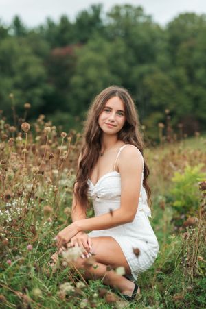 A young woman in a white summer dress sits in a wildflower meadow surrounded by nature and greenery.