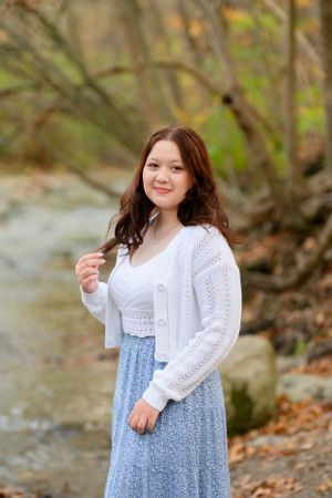 A person in a white knit cardigan and floral skirt stands near a creek on an autumn day.