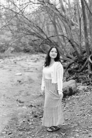 Black and white portrait of a smiling woman in a long skirt and cardigan standing on a forest path by a stream.