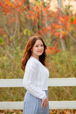 Person in white knit sweater and floral skirt poses beside white fence with vibrant autumn foliage in background.