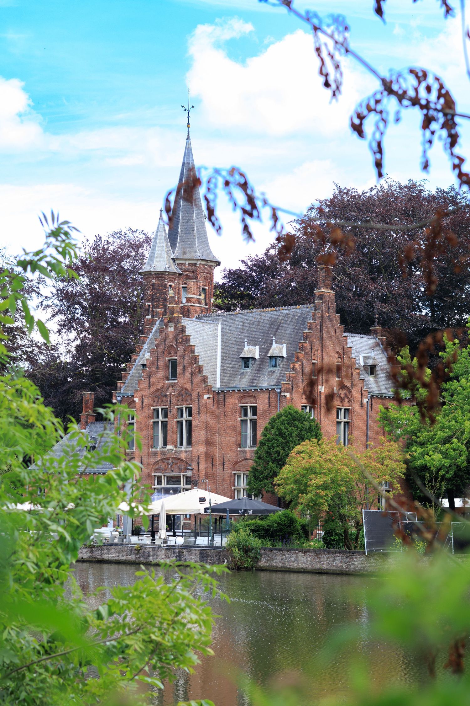 Medieval-style castle buildings with red brick towers situated beside a tranquil pond.