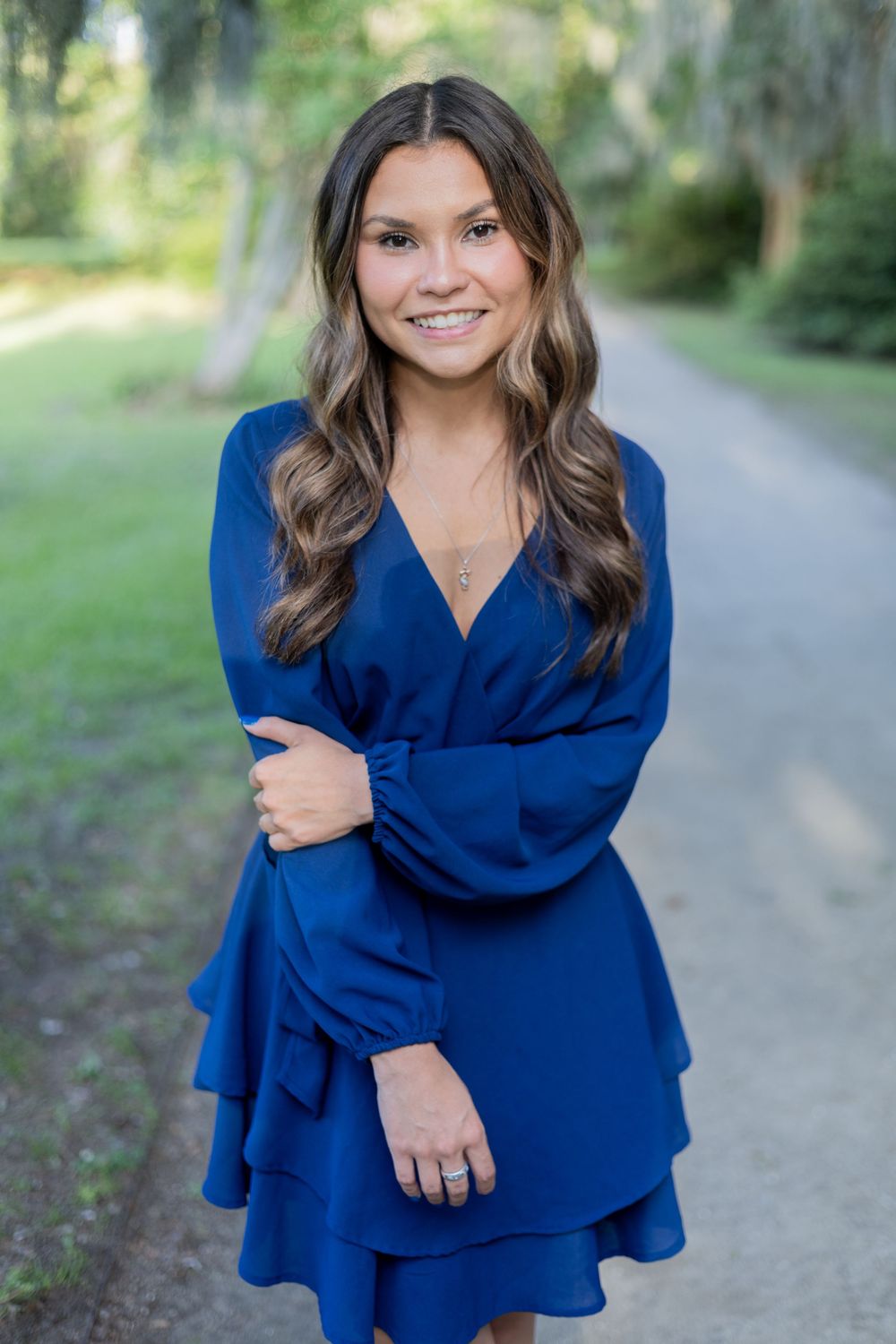 Person wearing a royal blue wrap dress with ruffle layers stands on a sunlit outdoor path surrounded by greenery.