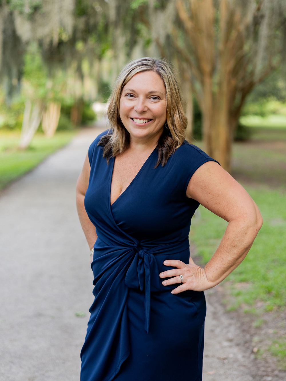 Woman in navy blue wrap dress poses confidently on tree-lined path in outdoor setting.