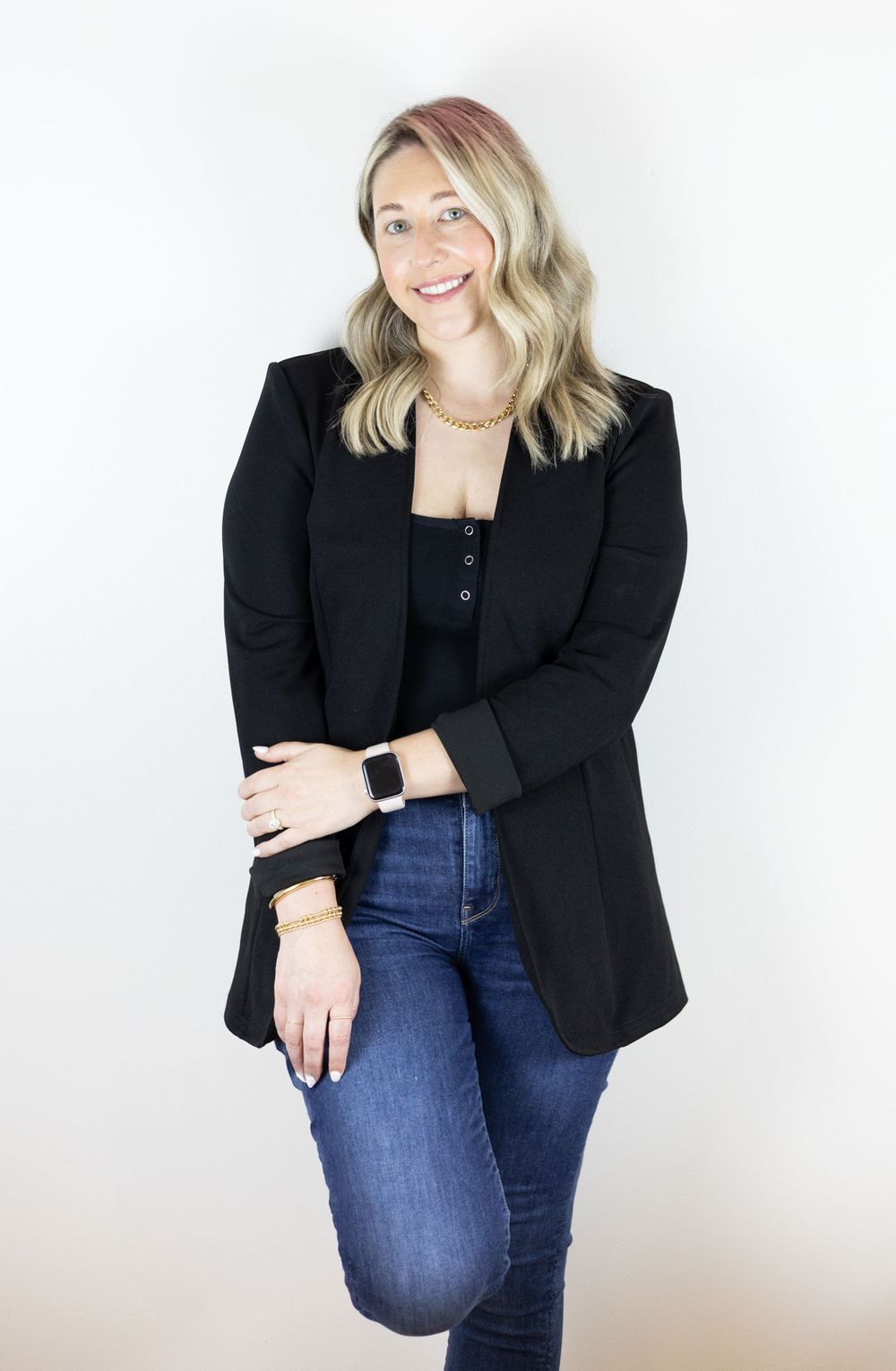 Professional studio portrait wearing a black blazer, gold necklace and blue jeans against a white background.