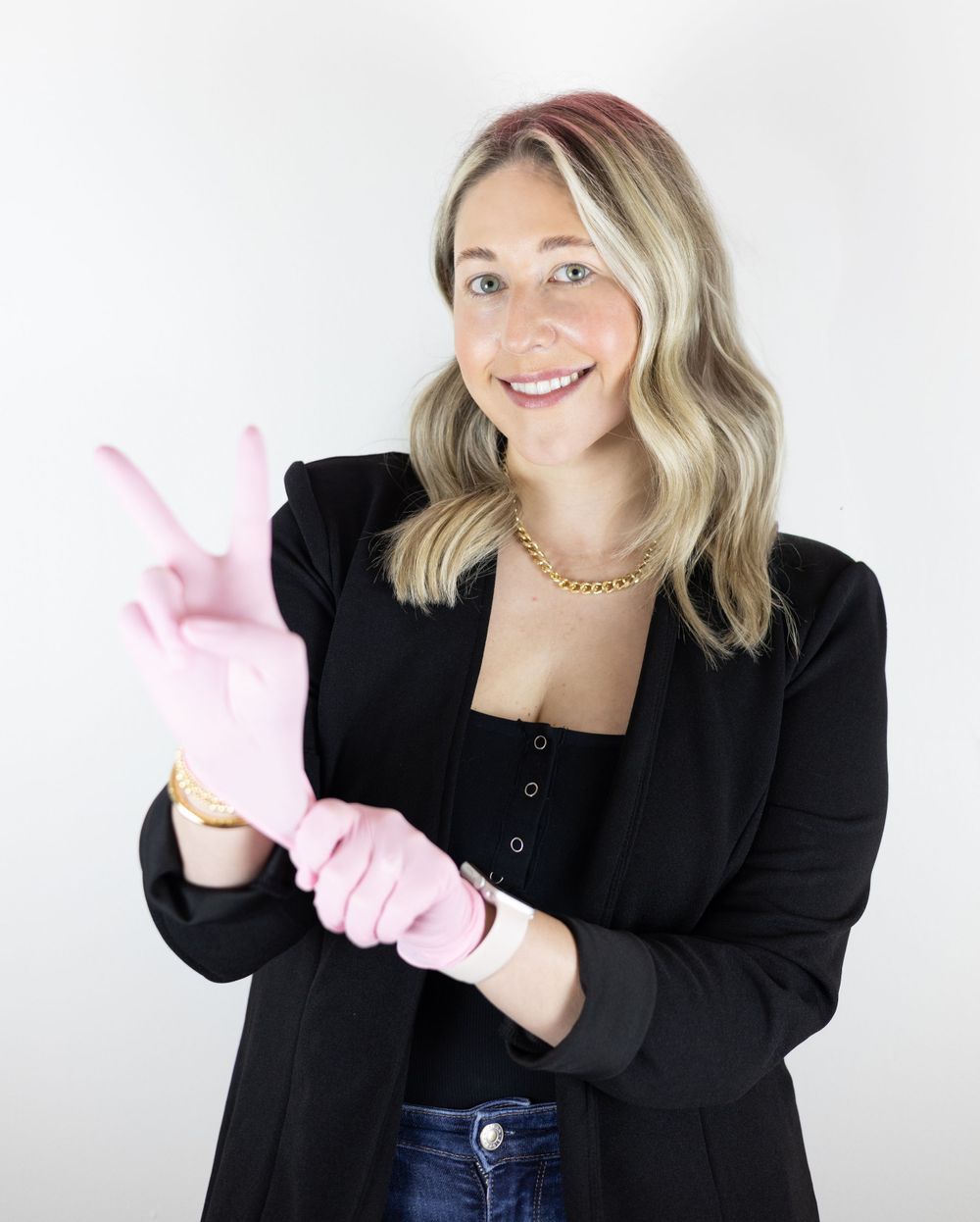 Professional wearing a black blazer and jeans adjusts a pink rubber glove while smiling against a white background.