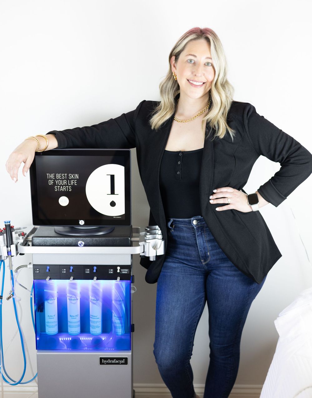 Professional standing beside a skincare treatment machine with blue LED lights and wearing black blazer and jeans.
