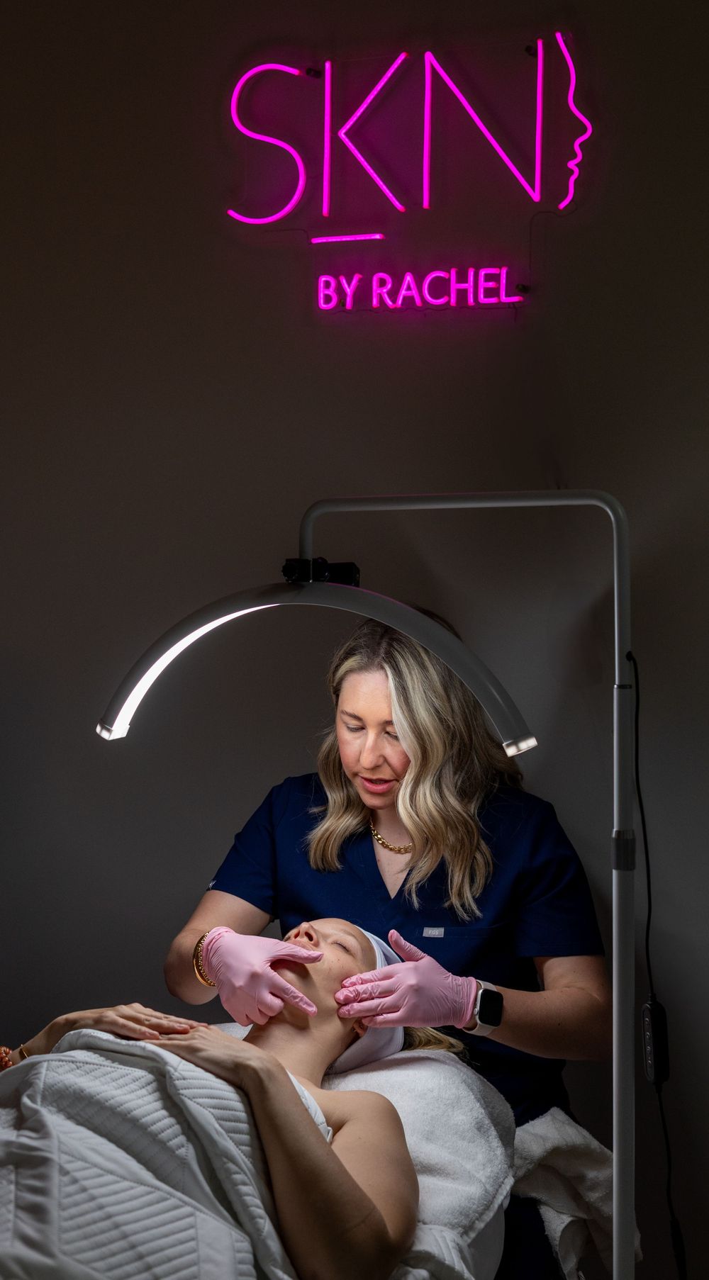 Esthetician performing a skincare treatment under a pink neon sign in a spa setting.