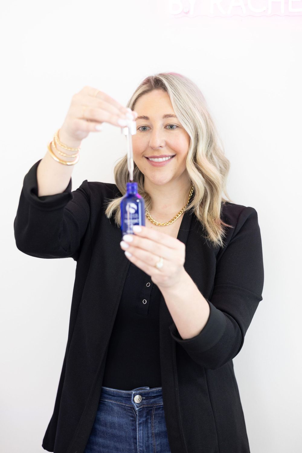 Professional woman in black blazer holding a small blue essential oil or serum bottle and dropper on light background.