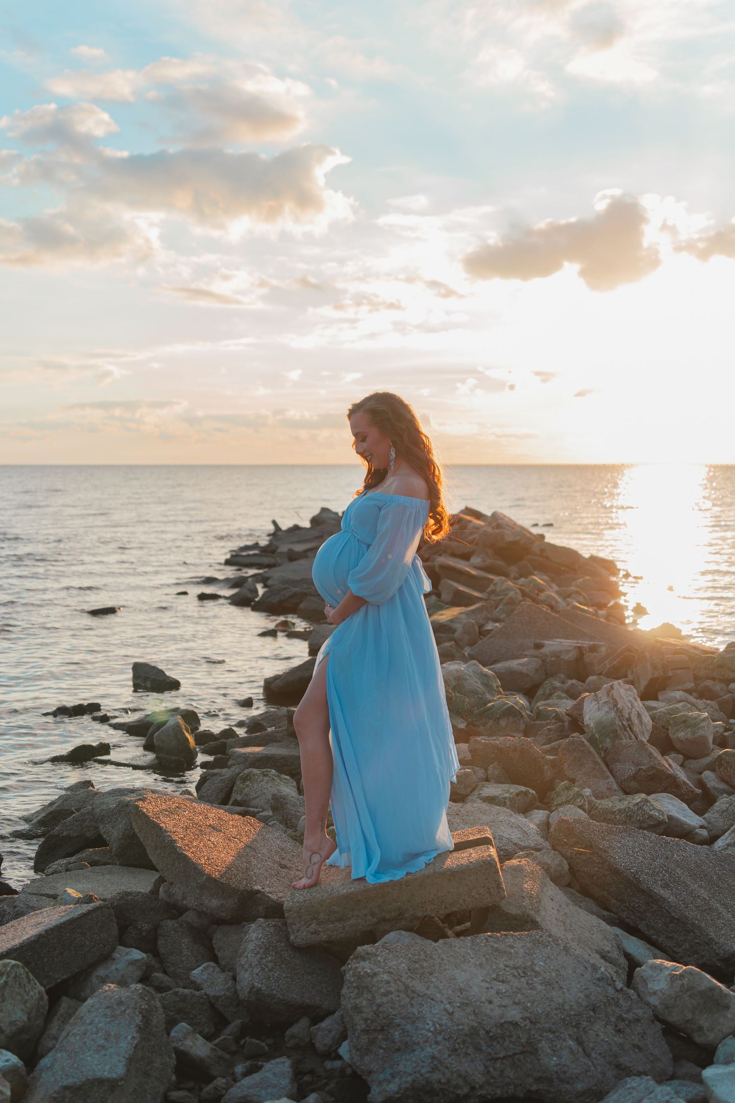 Silhouetted figure in flowing white dress stands on rocky beach at sunset with golden clouds and ocean waves in background.