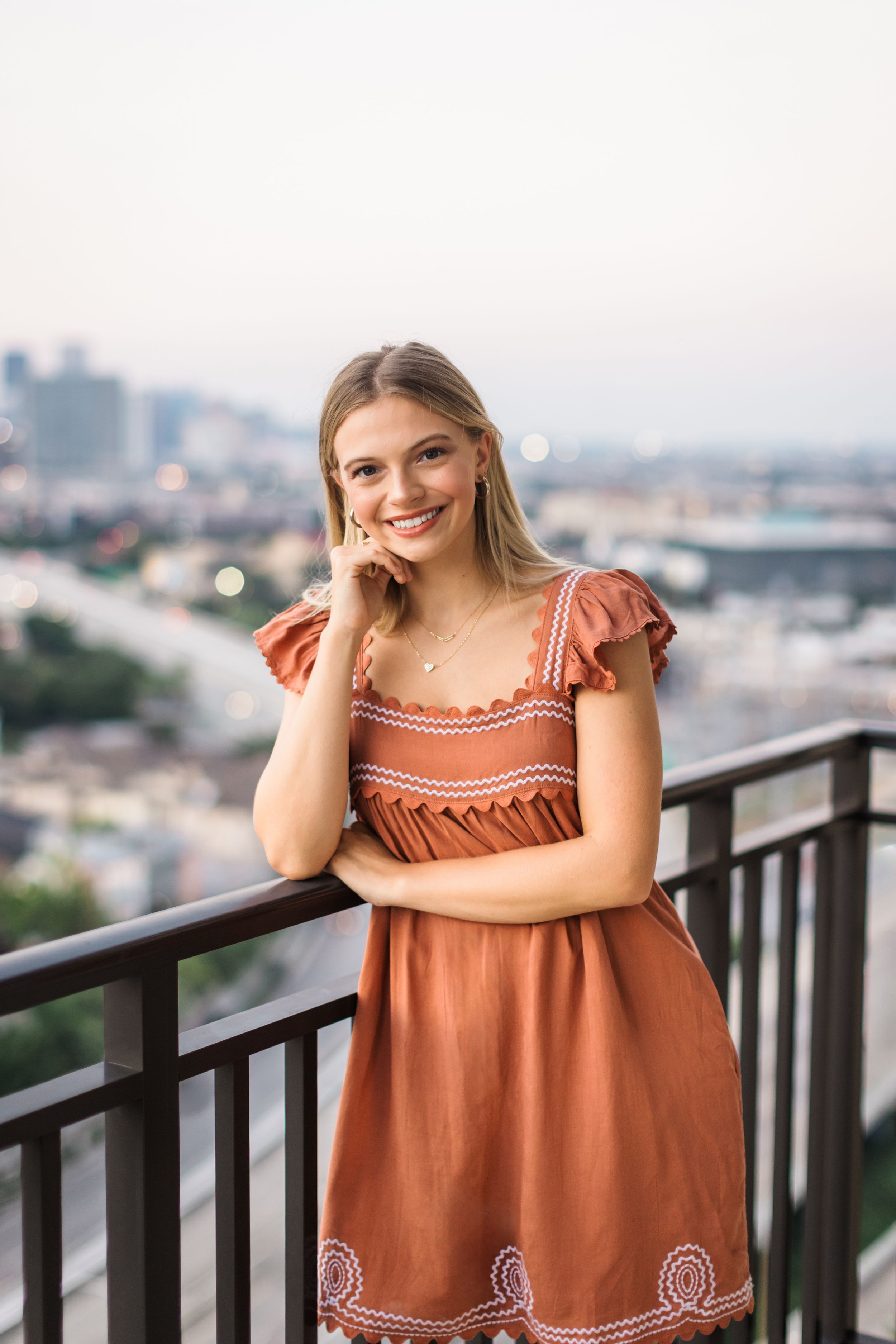 A person in a terracotta dress poses against metal railings on a balcony with city views.