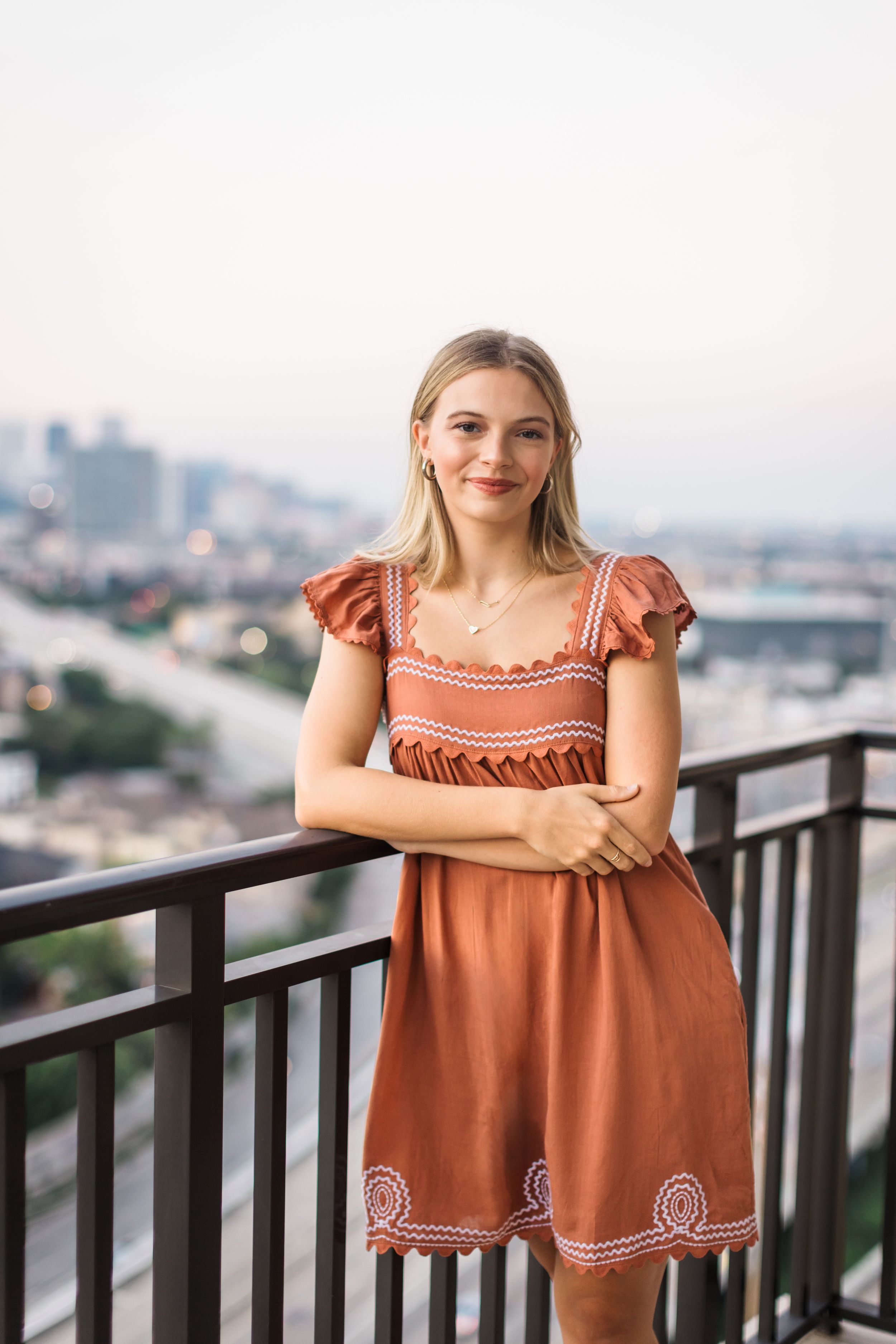A person wearing a rust-colored dress with embroidery details poses on an urban balcony at dusk.