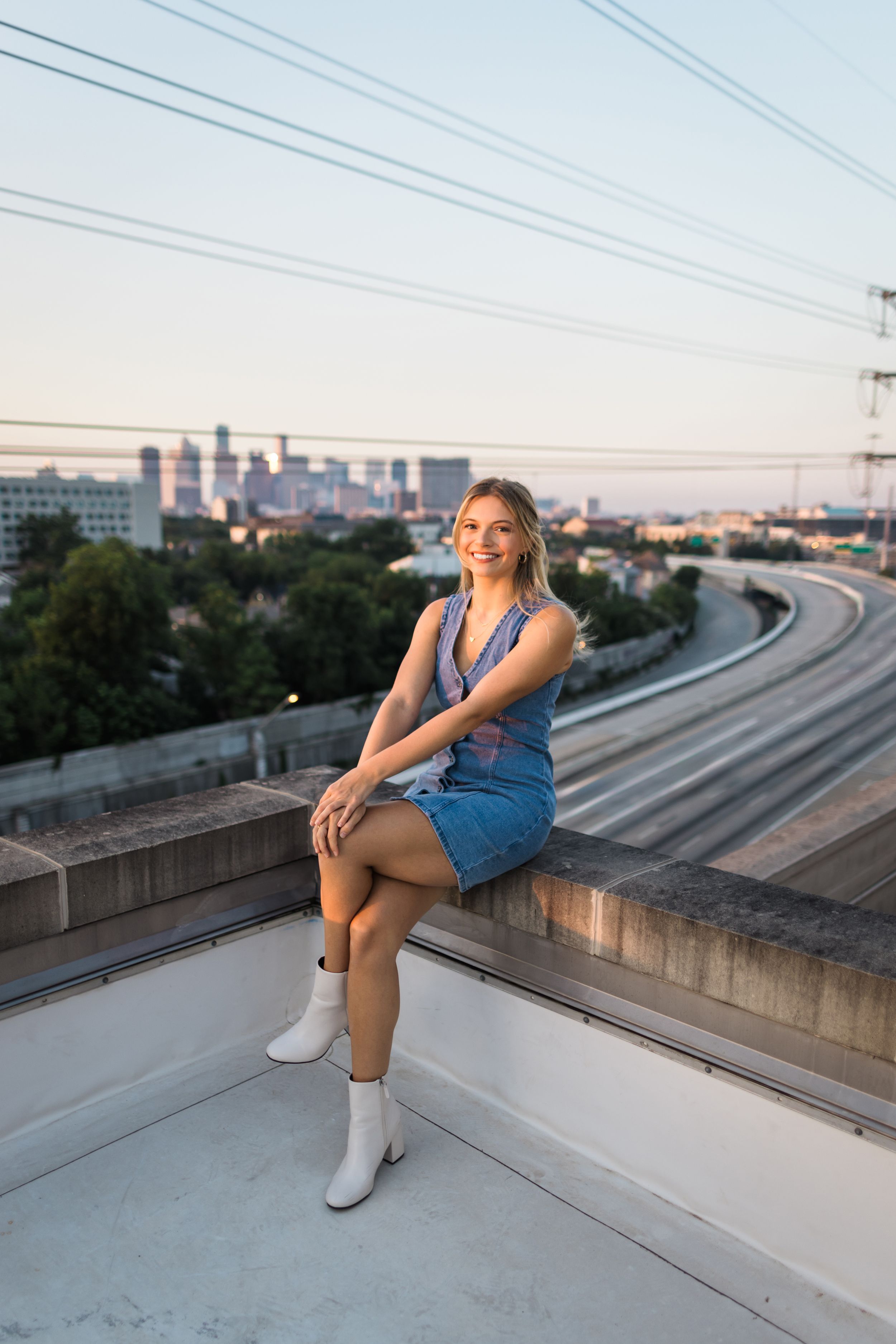 A person in a denim dress and white boots poses on a concrete ledge overlooking train tracks and city skyline at sunset.