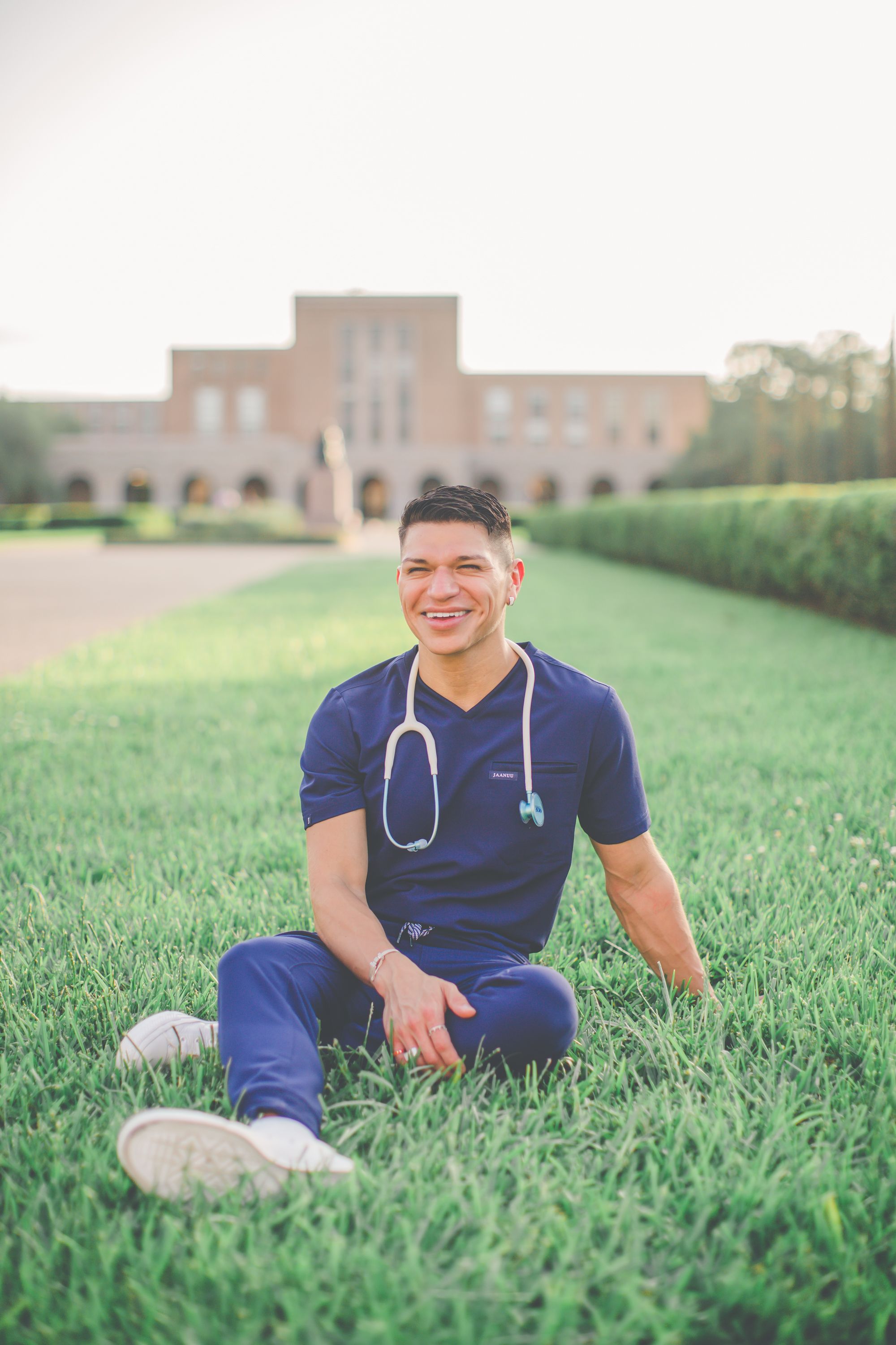 Healthcare worker in navy scrubs relaxes on grass with university building in background.