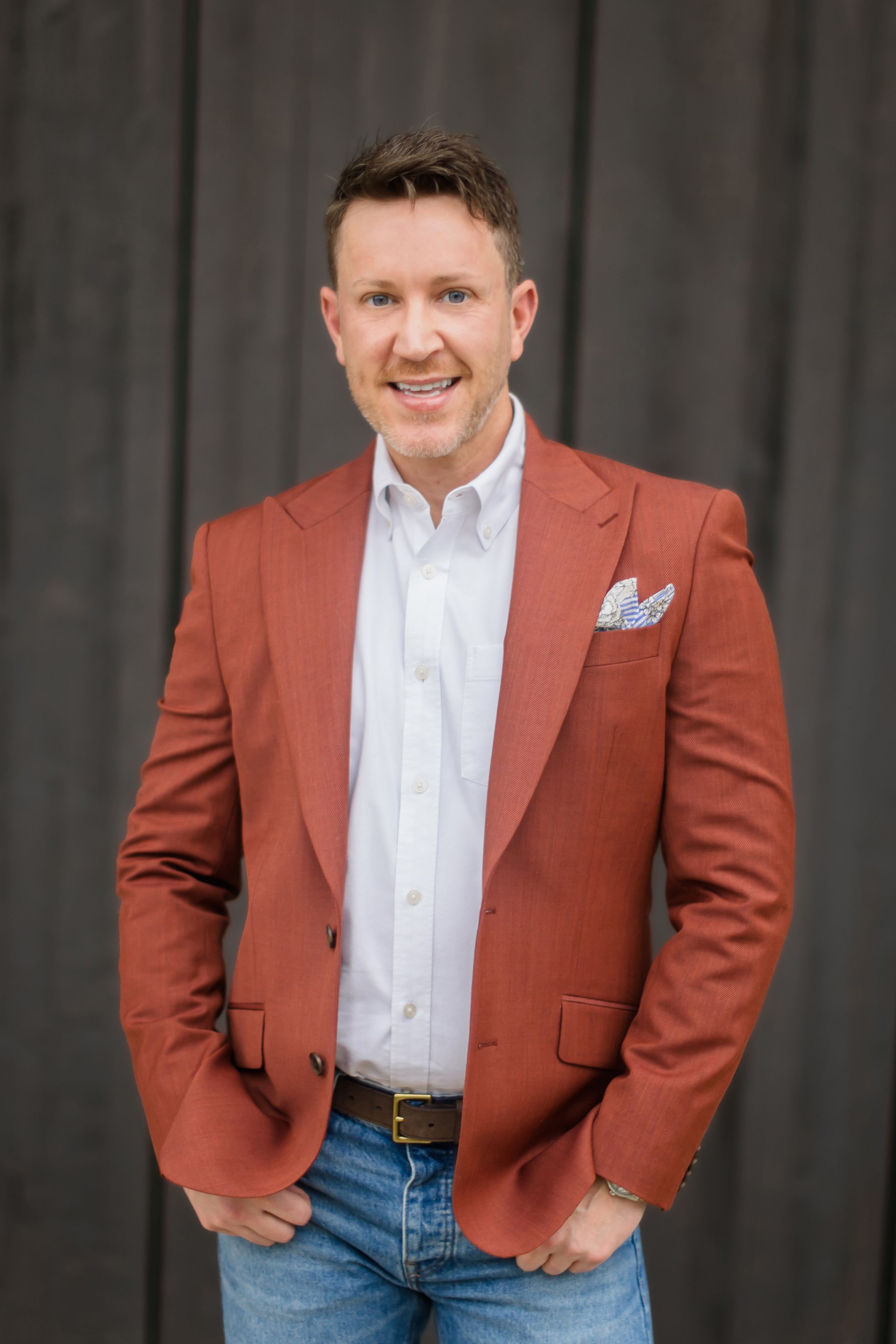 Professional headshots of person in rusty brown blazer and white shirt against dark backdrop.