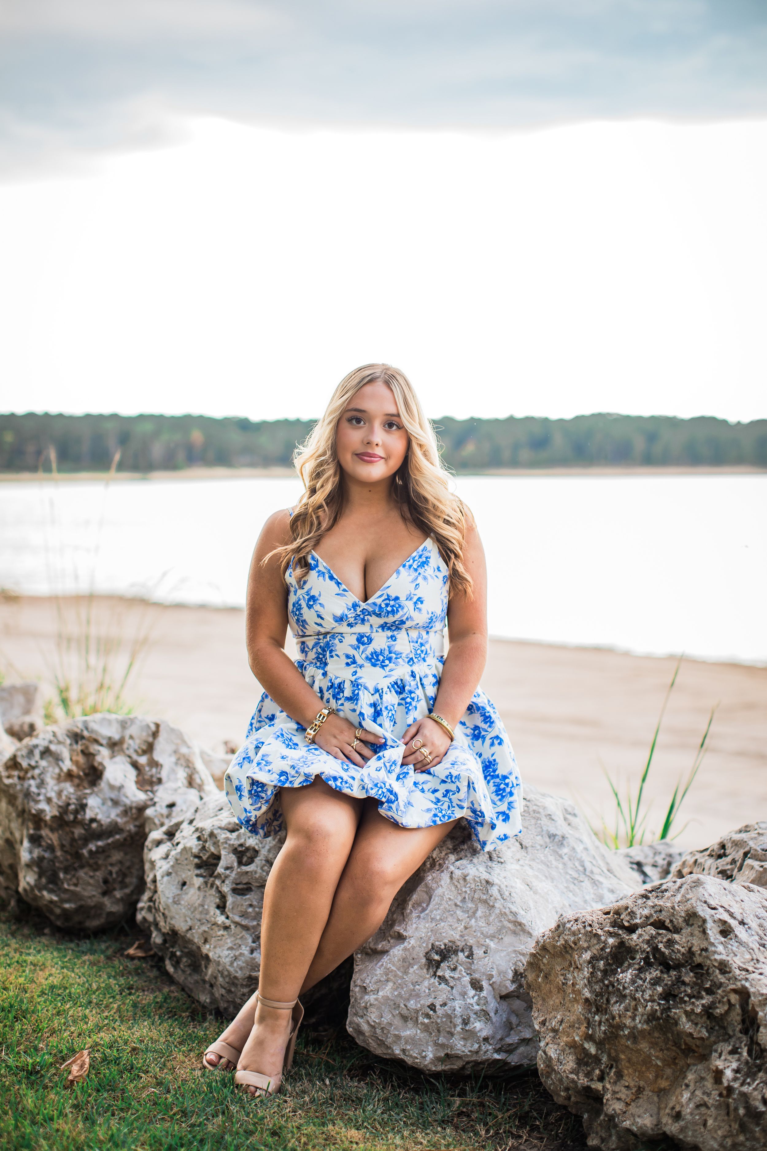 A person in a blue and white floral dress sits on large rocks by a serene lake on a cloudy day.