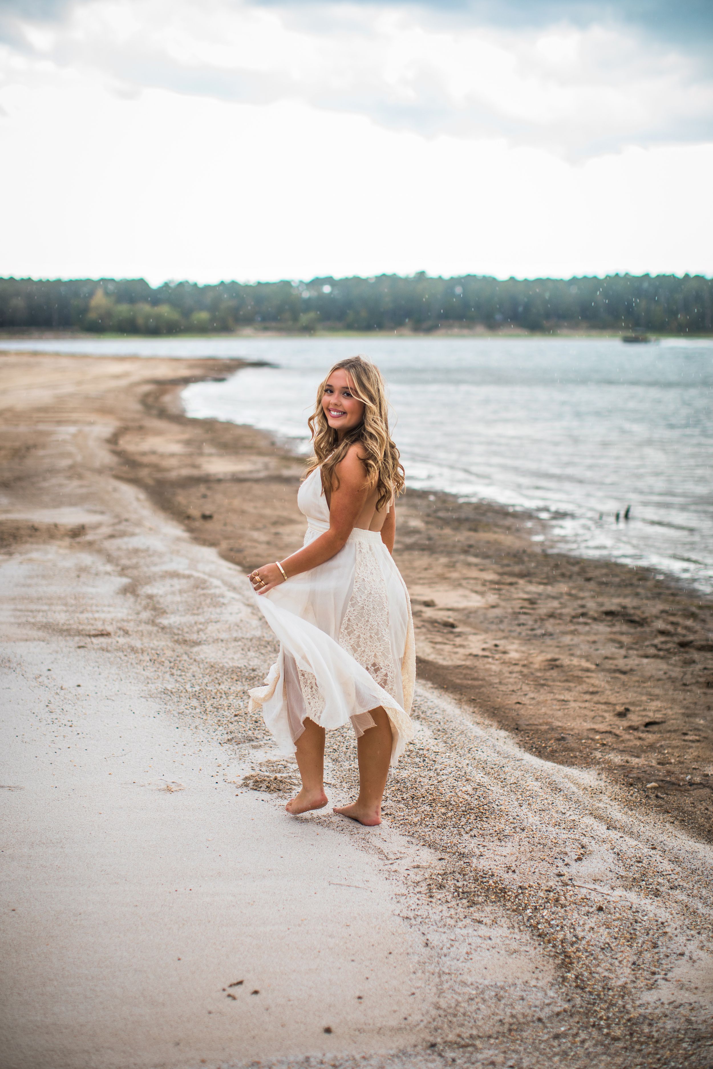 A person in a flowing white dress crouches down on a sandy beach shoreline near calm waters and forest.