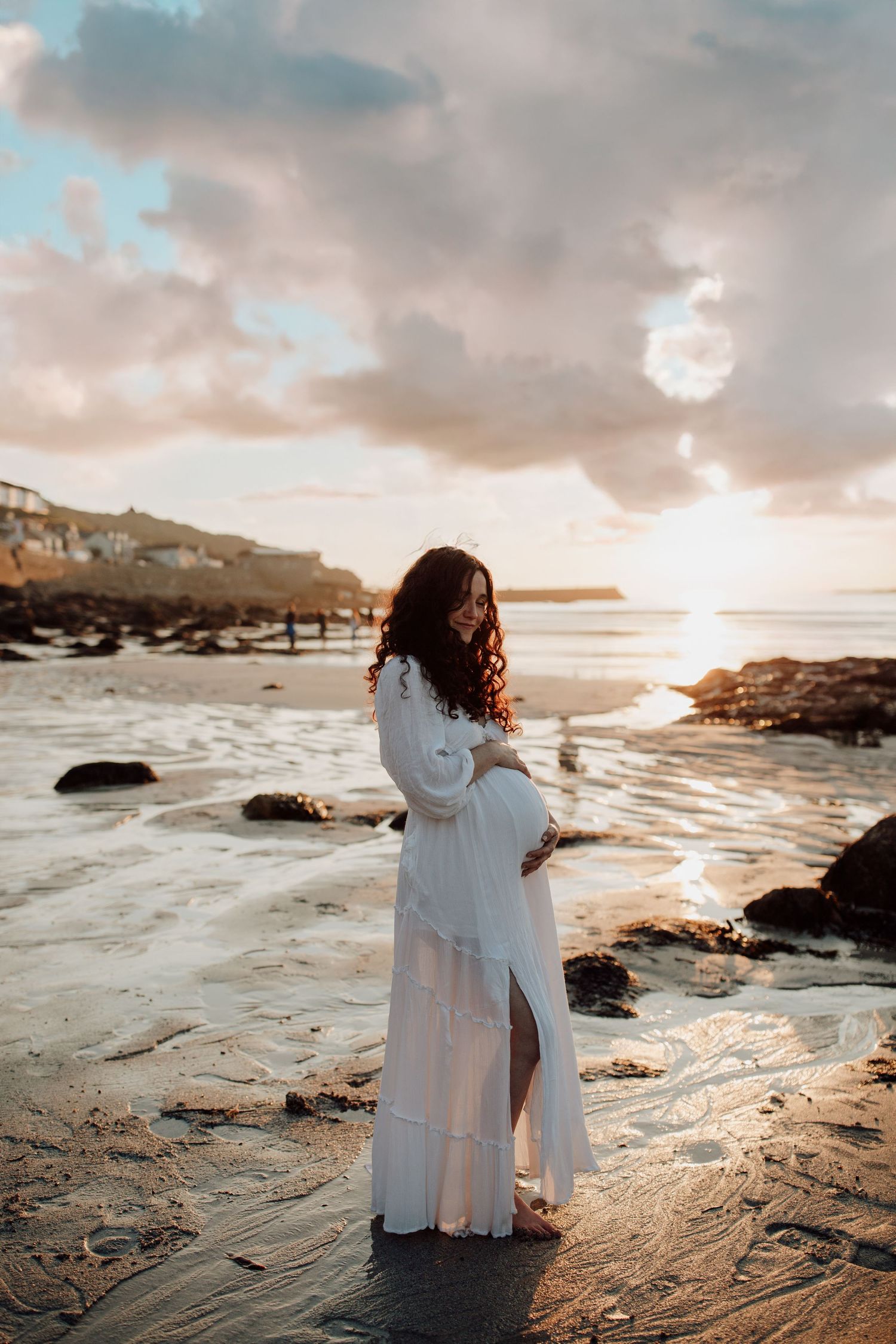 A figure in a flowing white dress stands on a rocky beach at sunset, with waves crashing against the shore and cliffs in background.