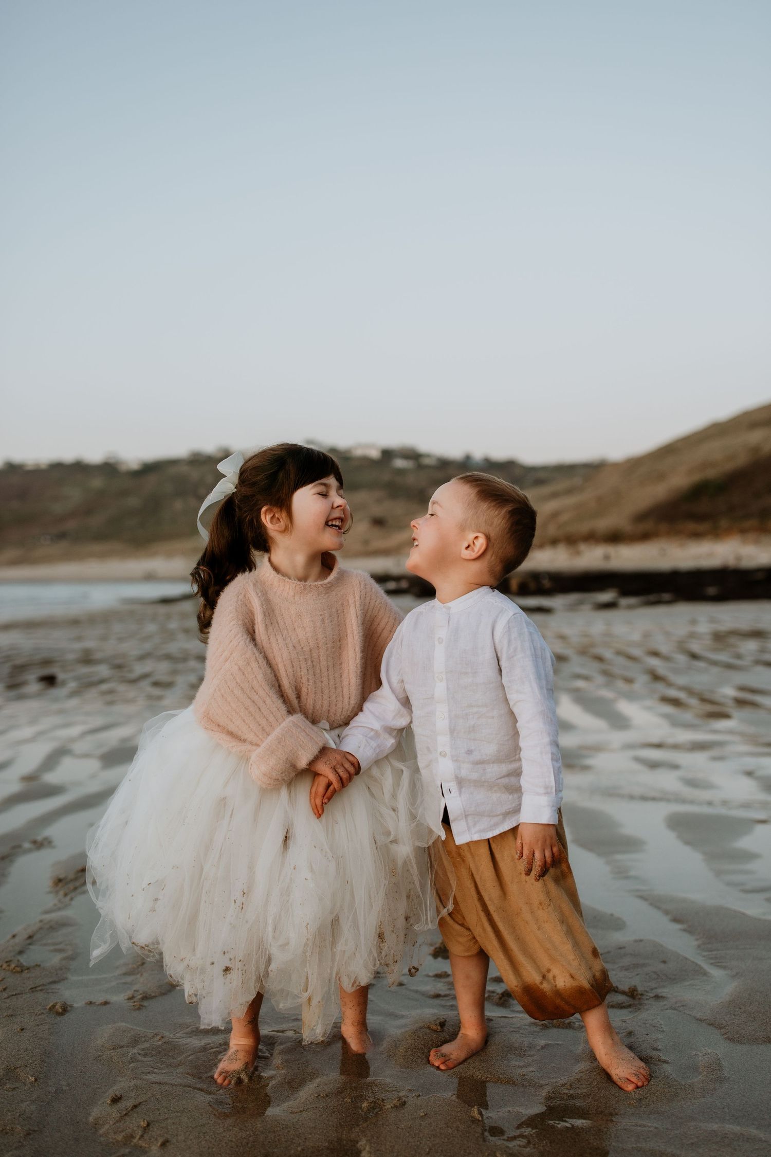 Two young children laughing and holding hands while standing barefoot on a beach at sunset.