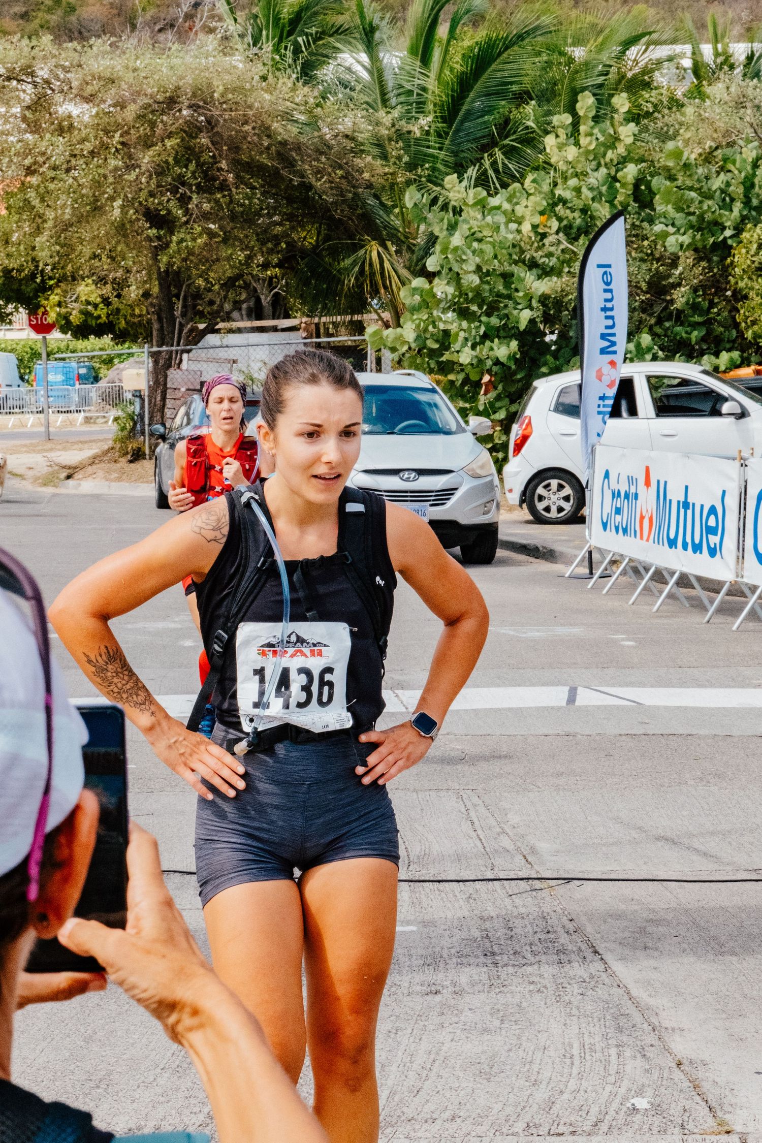 Athletic runner in black tank top and shorts catches breath after race near event barriers and palm trees on sunny day.