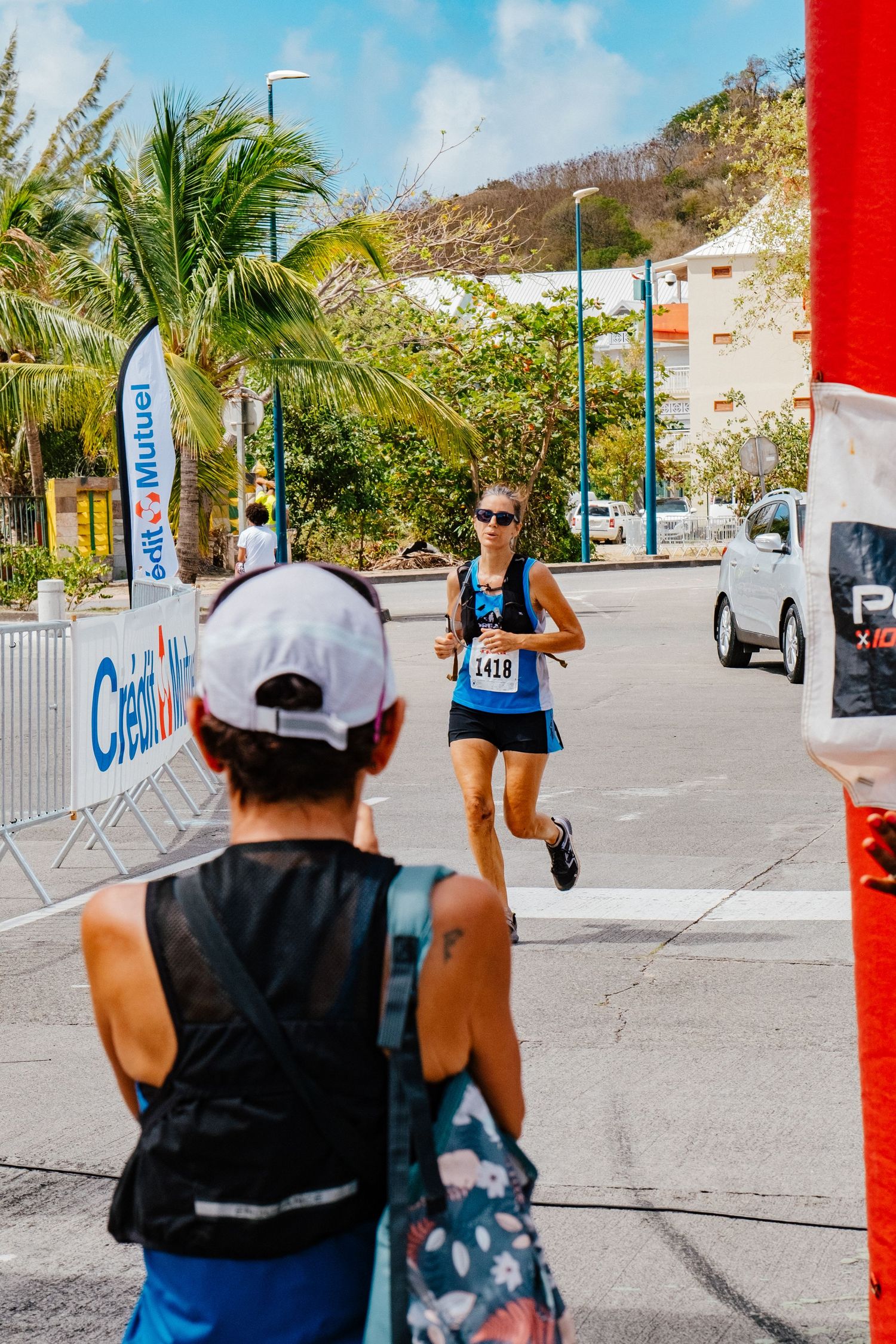 A runner in blue during a sunny race event with palm trees and banners along a barricaded street course.