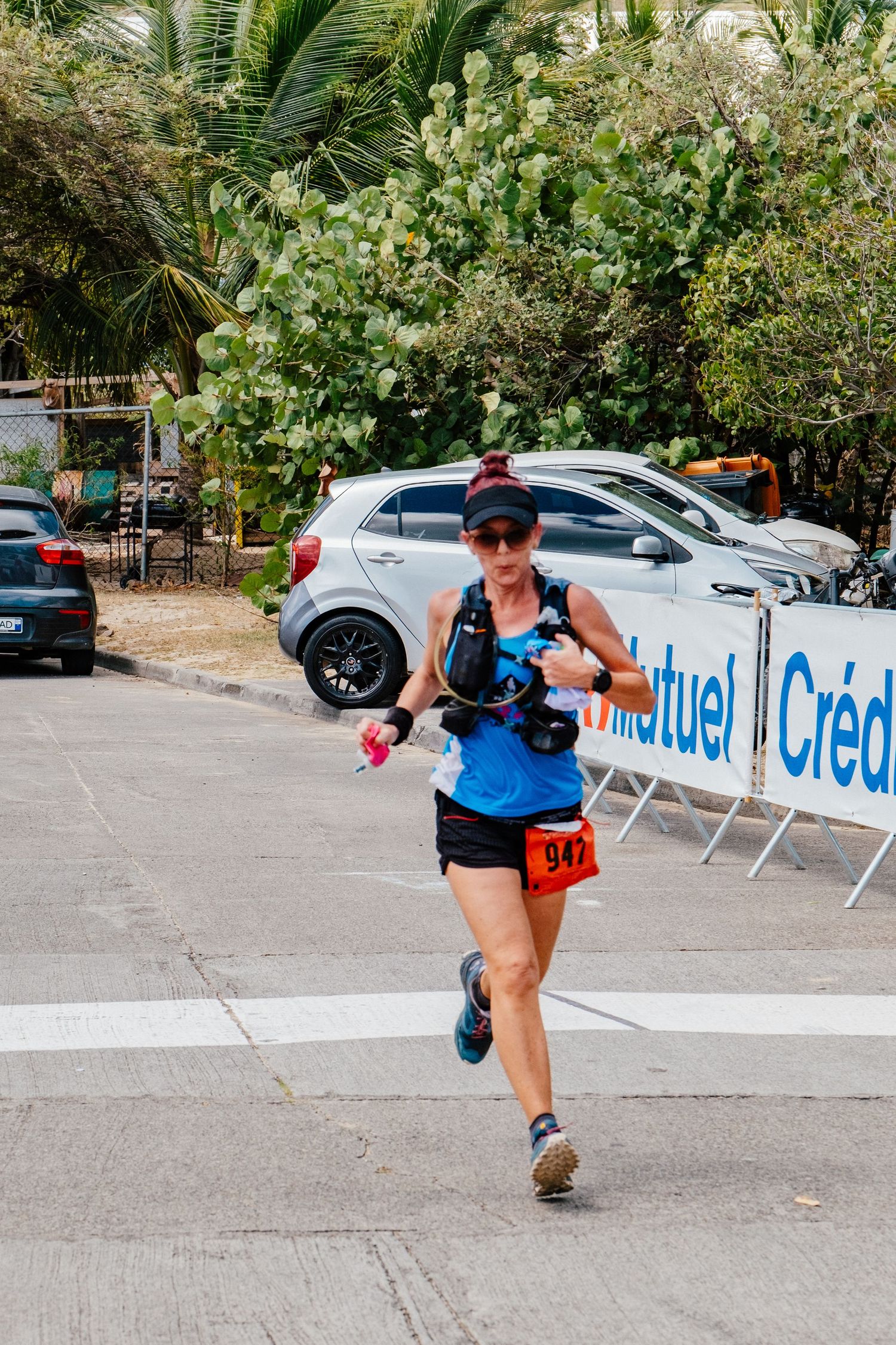 Runner in blue tank top and black shorts competing in a race with palm trees and parked cars in the background.