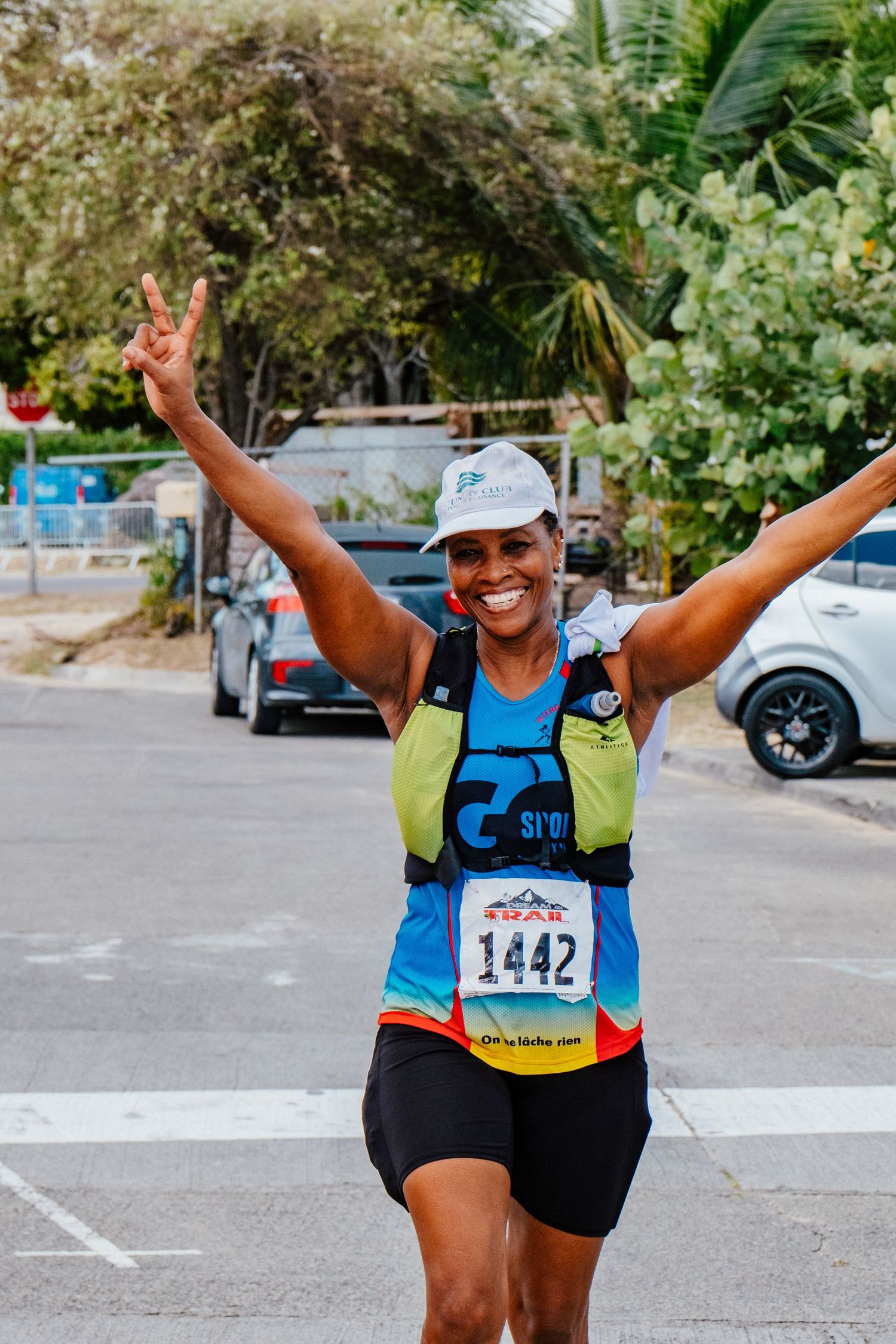 Race participant wearing bib number 1442 celebrates with raised arms and peace sign during outdoor running event.