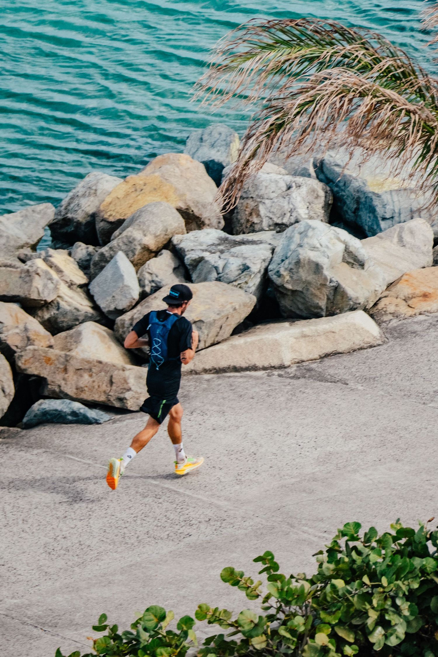 A runner jogs along a coastal path with large rocks and turquoise water in the background beneath palm fronds.