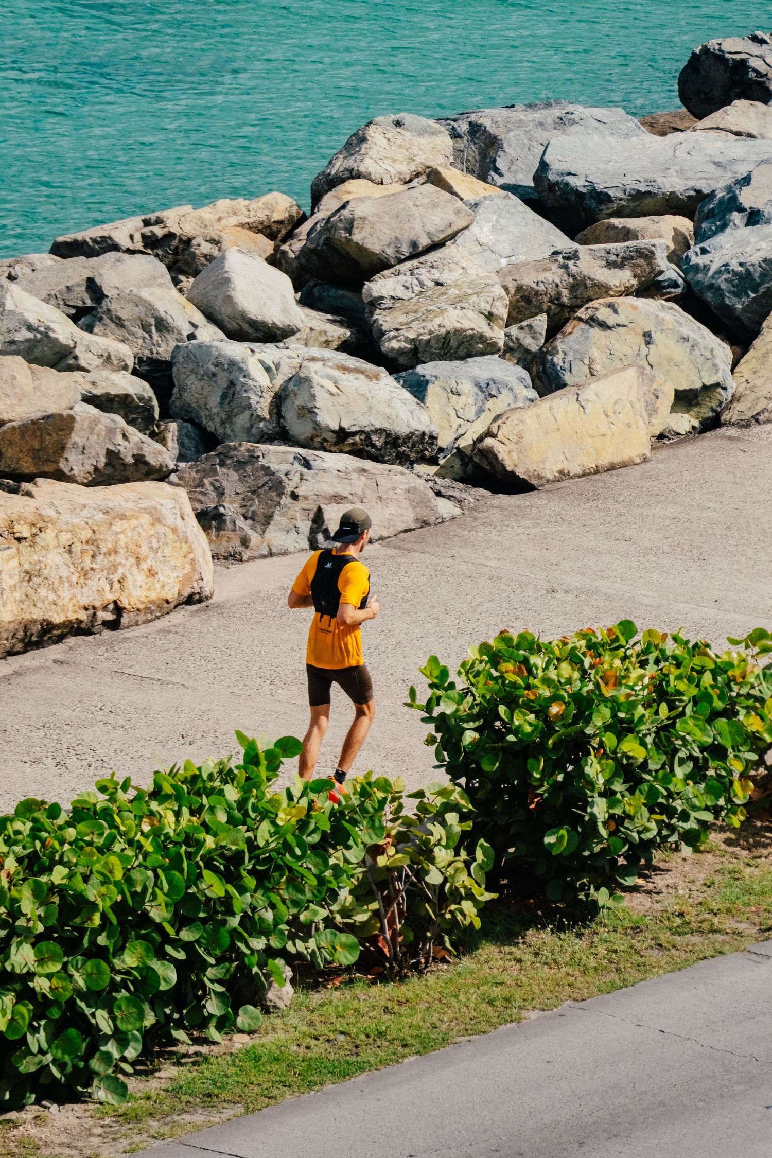 Person in yellow backpack walking along coastal path with turquoise water and large rocks beside tropical foliage.