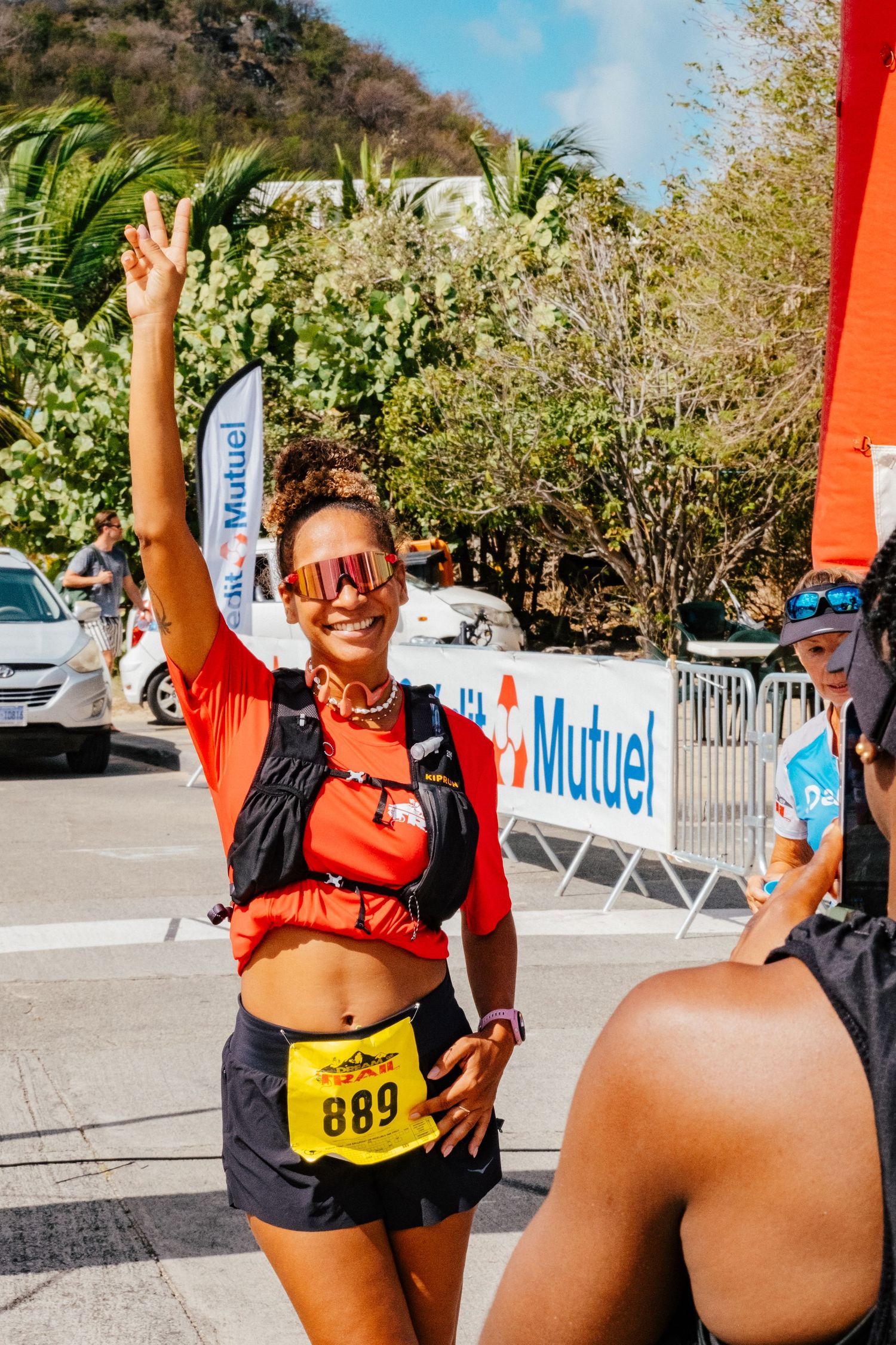 Runner in orange top and dark shorts flashes peace sign at outdoor race event with palm trees in background.