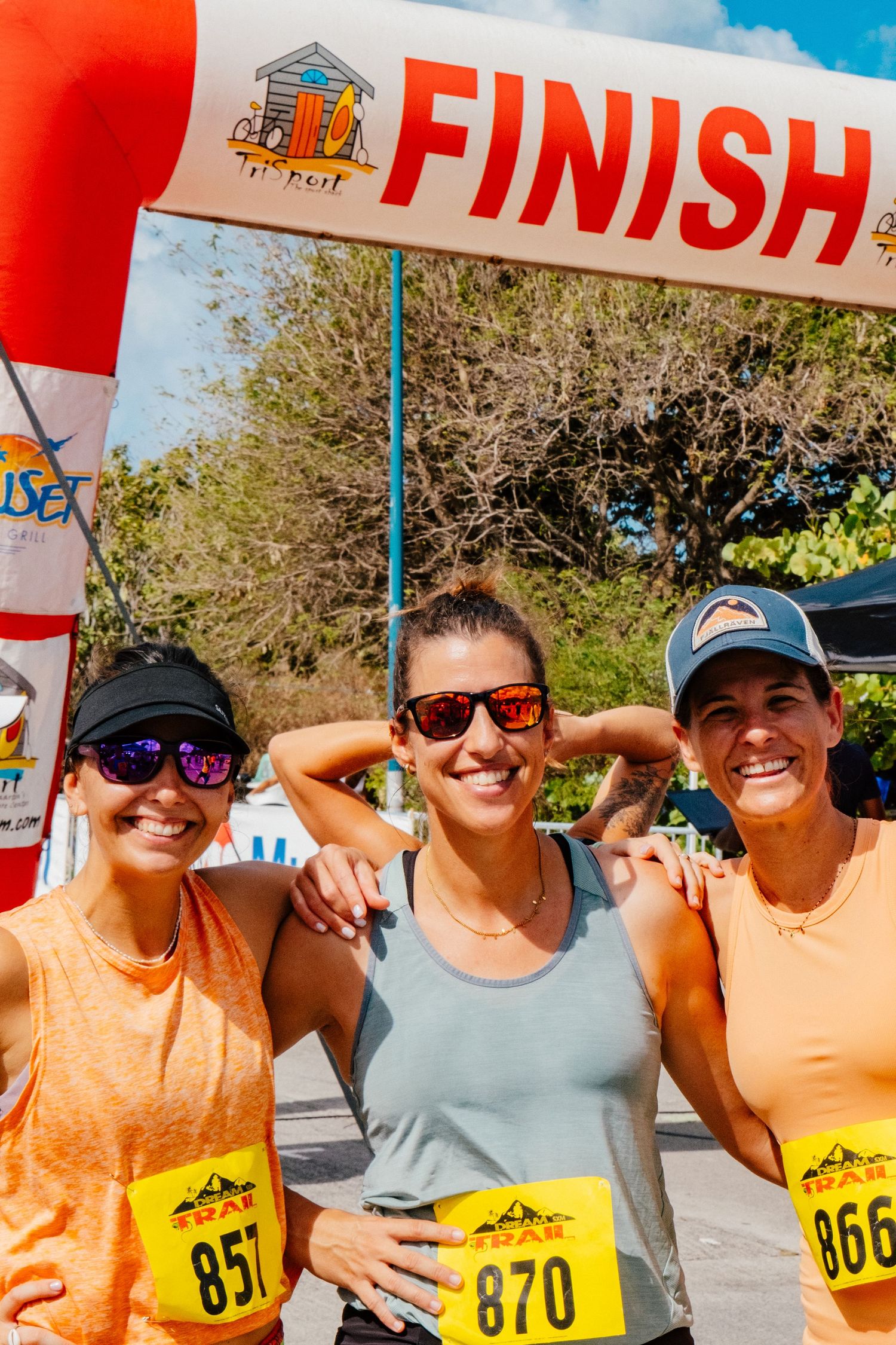 Three smiling runners pose together under a finish line banner at an outdoor race event wearing numbered bibs in sunny weather.