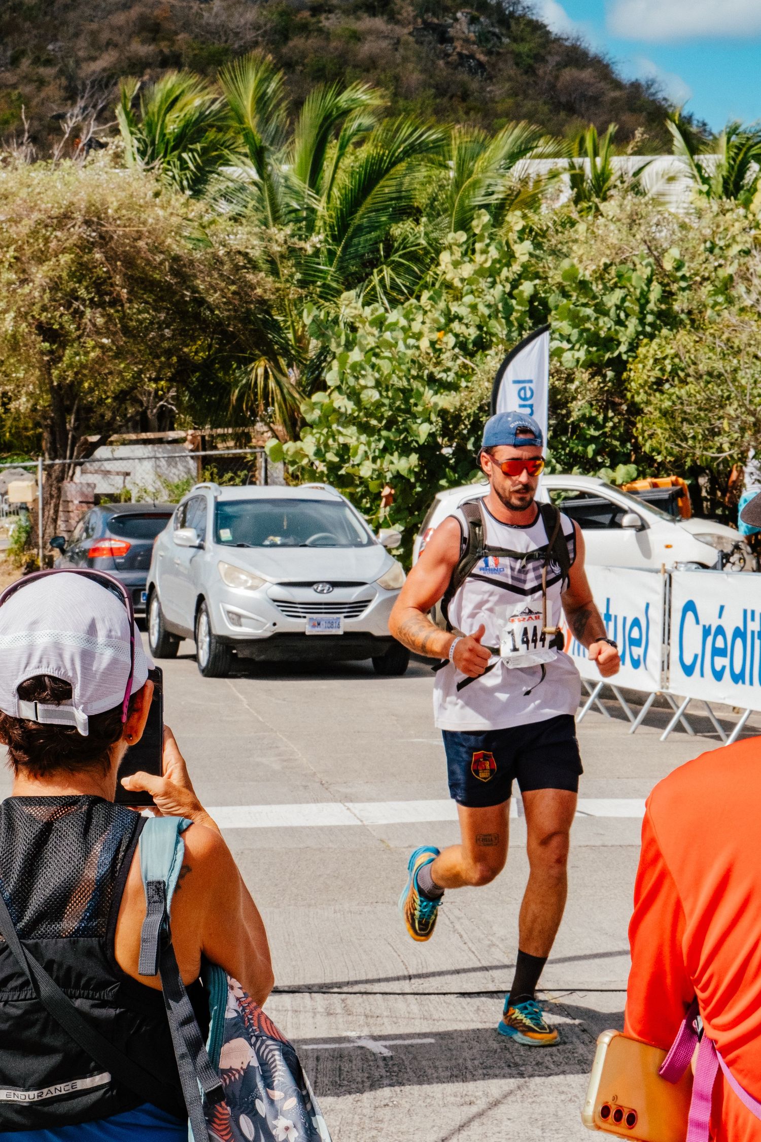 Runner in blue cap and tank top racing along tropical course with palm trees and vehicles in background during event.
