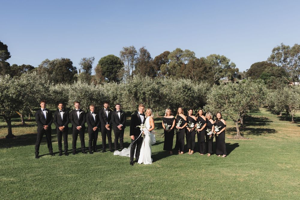 A wedding party in black and white attire poses on a lush green lawn surrounded by trees at an outdoor venue.