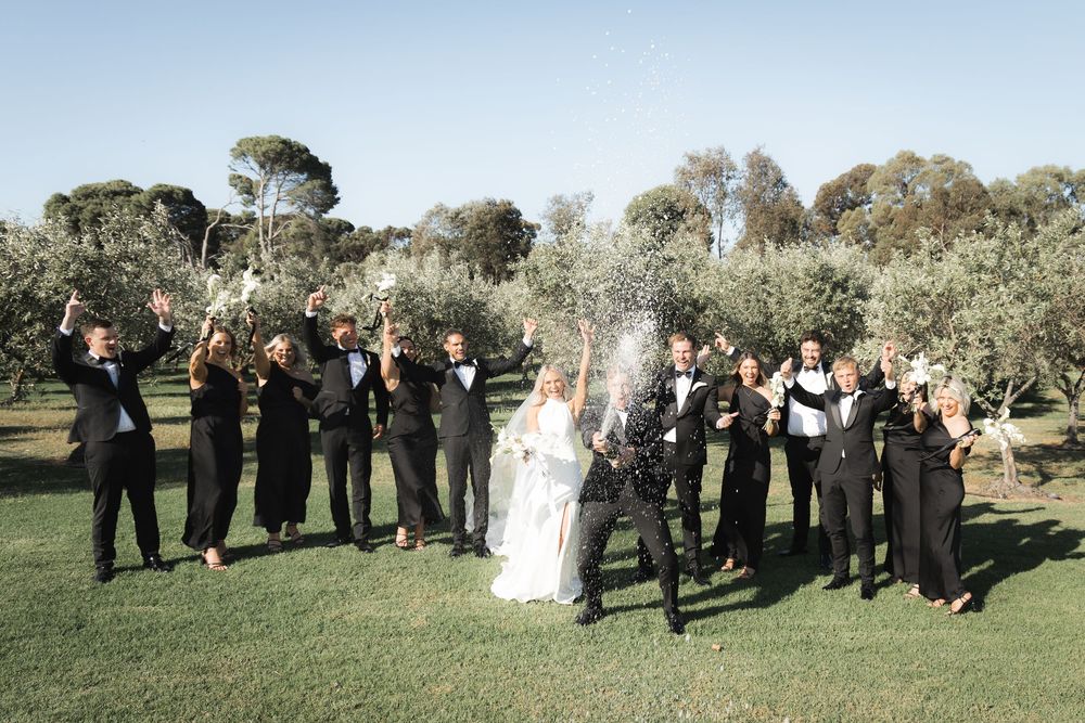 Wedding party celebrating with champagne spray in an outdoor garden setting surrounded by olive trees.