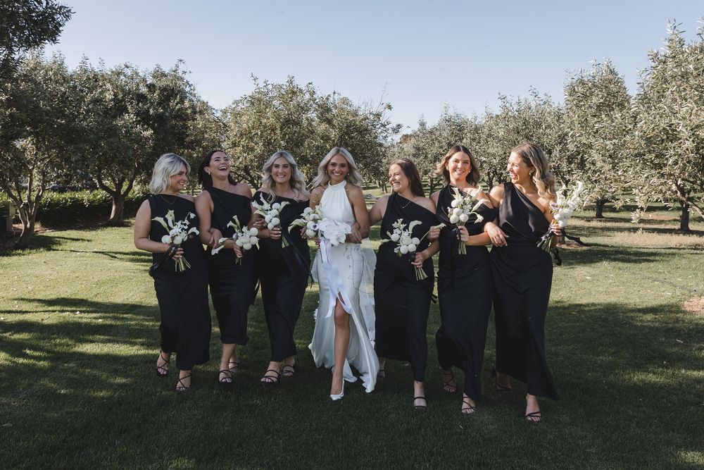 Wedding party in black dresses and white gown pose together holding bouquets in an orchard on sunny day.