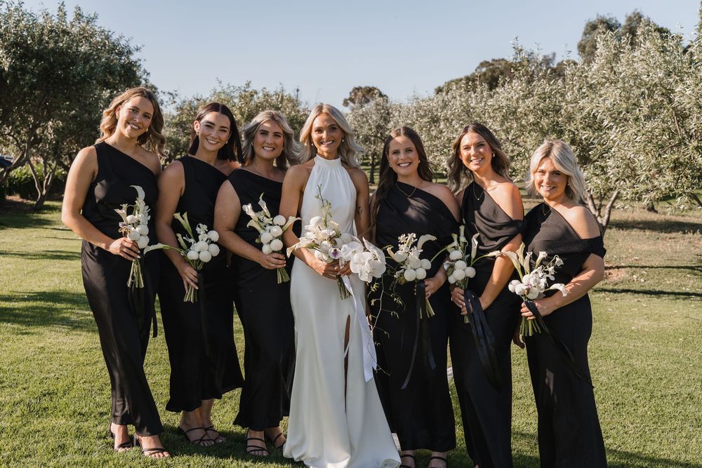 Wedding party in black dresses and white gown poses together in sunny orchard setting.
