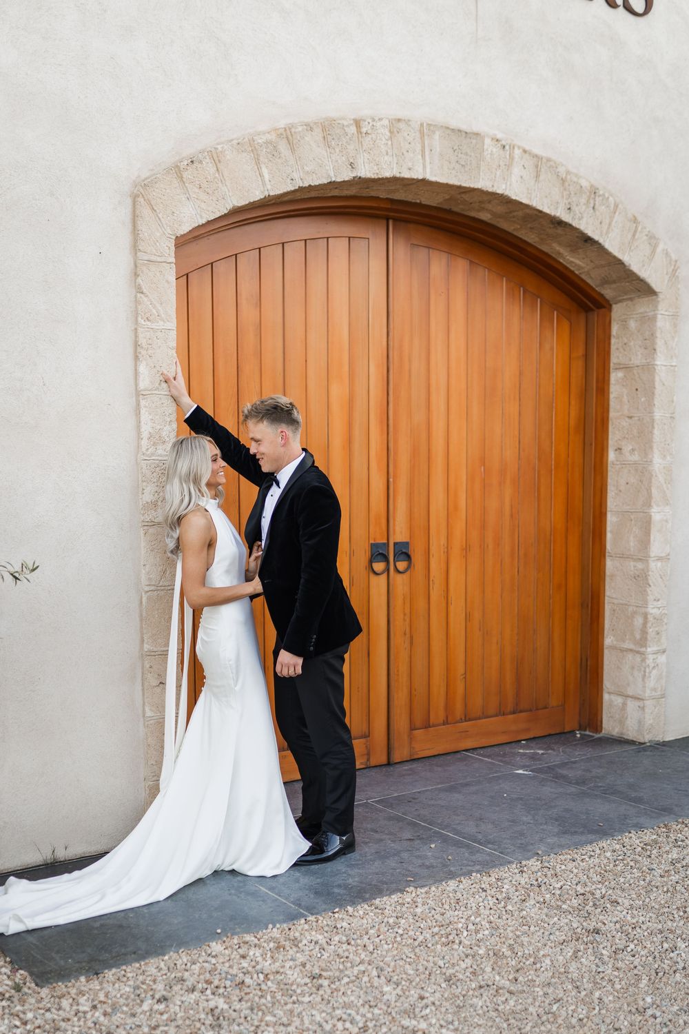 A couple in wedding attire embraces by a large wooden arched door with stone trim and white stucco walls.
