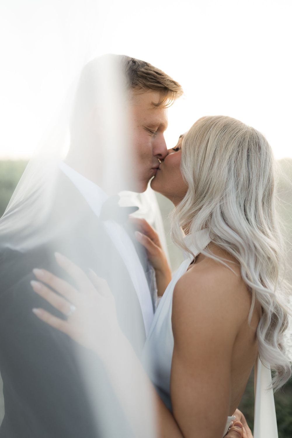 A romantic wedding couple shares an intimate kiss in soft natural light with dreamy white veil effect.