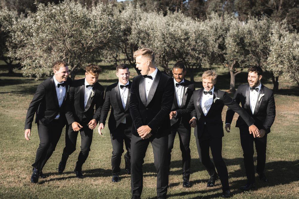 A group of men in black tuxedos and bow ties stand together outdoors in front of olive trees on a sunny day.
