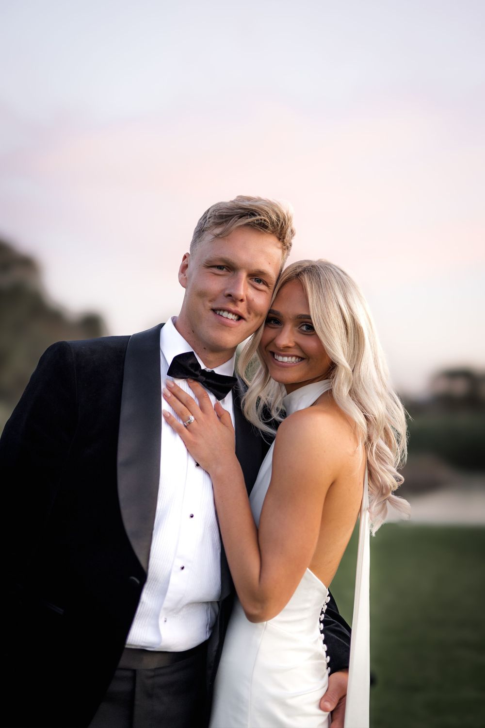 Elegant couple in formal wedding attire share romantic outdoor moment during sunset with the bride showing engagement ring.