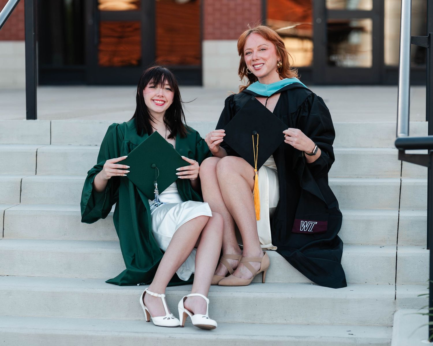 Two graduates in green and black robes sit on concrete steps celebrating their achievement.