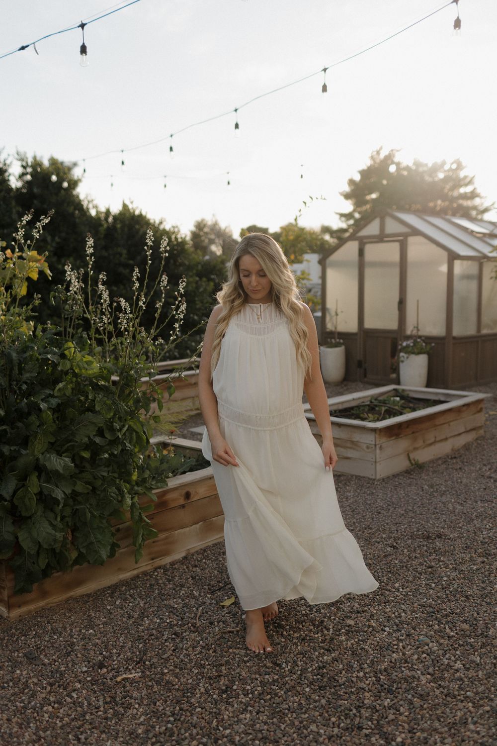 A flowing white dress catches the evening light in a garden setting with string lights and a greenhouse.
