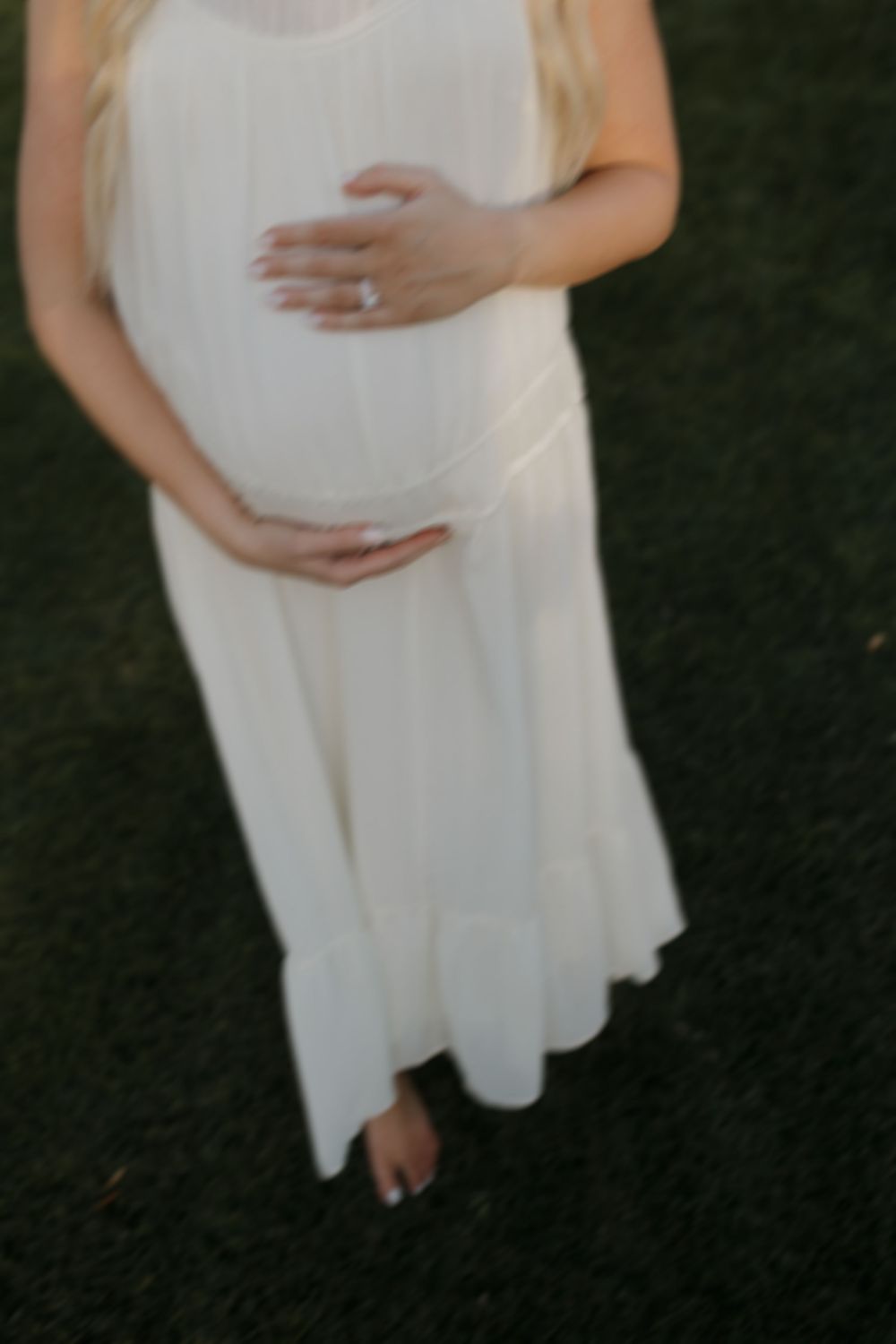 A gentle silhouette in a flowing white dress tenderly cradles a baby bump in an outdoor setting.