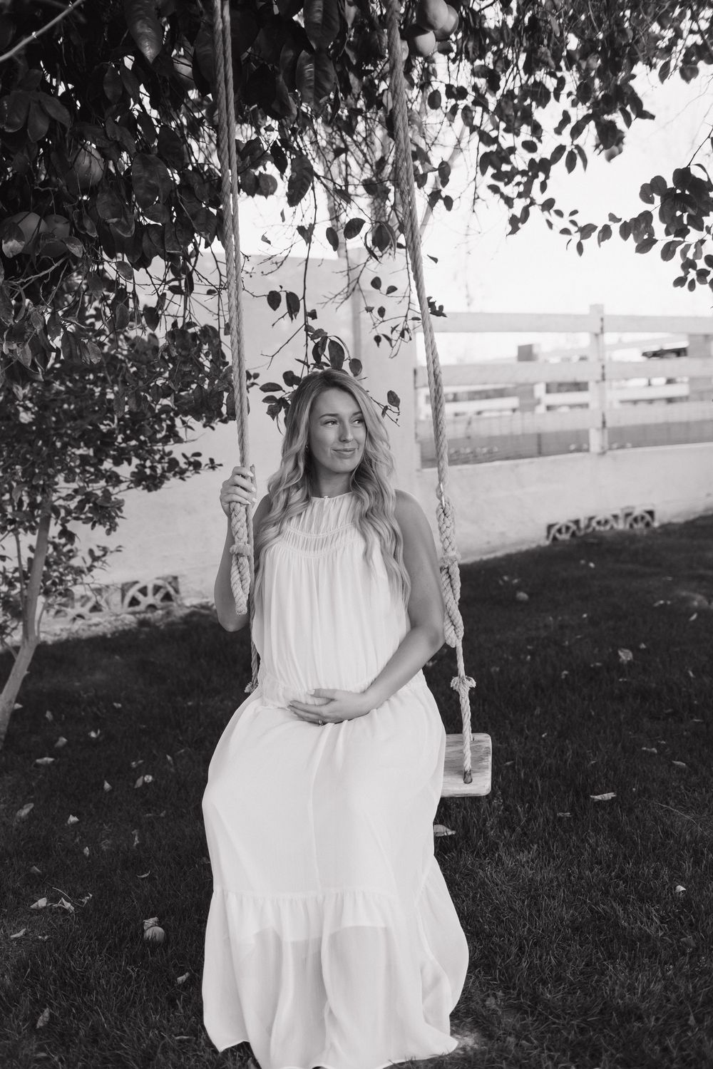 A black and white photo captures a peaceful moment of an expectant mother sitting on a wooden swing in a flowing dress.
