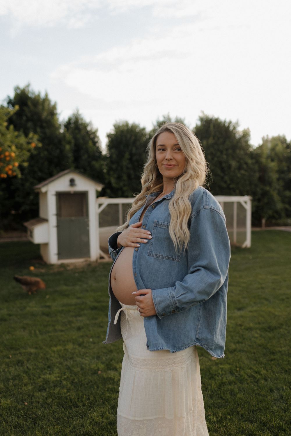 Expectant person in a white dress and denim jacket stands in a grassy field with white buildings in the background.