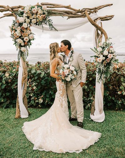 Rustic driftwood wedding first kiss arch decorated with peach roses and greenery frames a couple kissing by the ocean.
